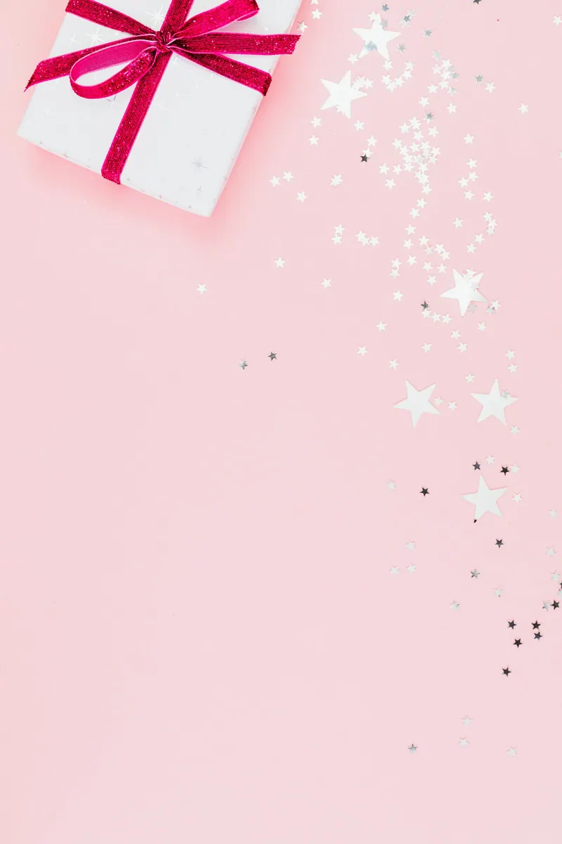 Top view of a white gift box with pink ribbon and silver star confetti on a pink background
