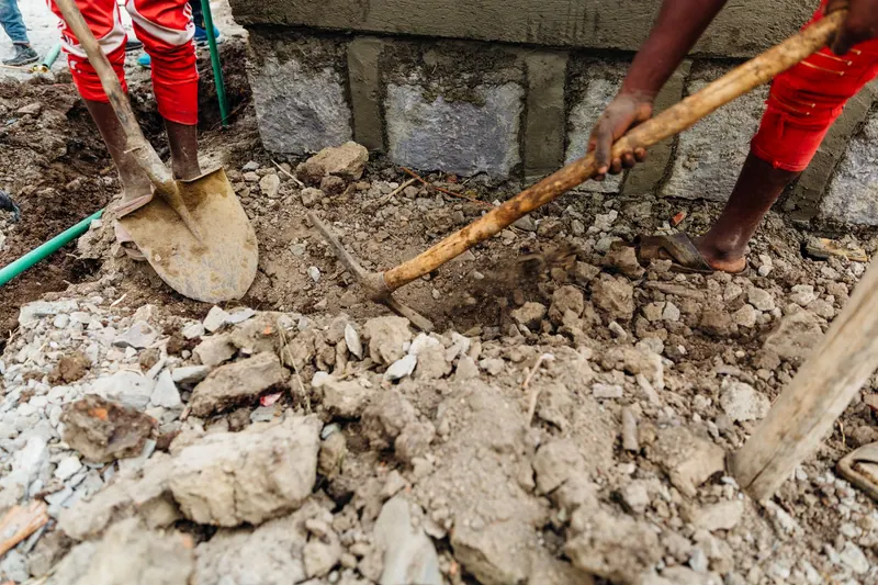 Workers using shovel and pickaxe during construction dig, showing manual labor on rocky ground