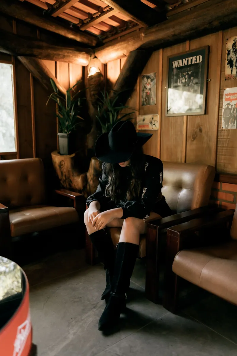 A cowgirl in a rustic wooden room with Western decorations, plants, and a wanted poster.